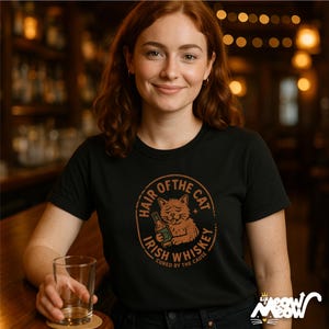 以下が含まれることがあります： 黒いTシャツ。円形のグラフィックには、ウイスキーのボトルを持った猫が描かれており、テキストには「Hair of the Cat, Irish Whiskey, Cured by the Cause」と書かれています。女性がそのシャツを着て、グラスを持っています。