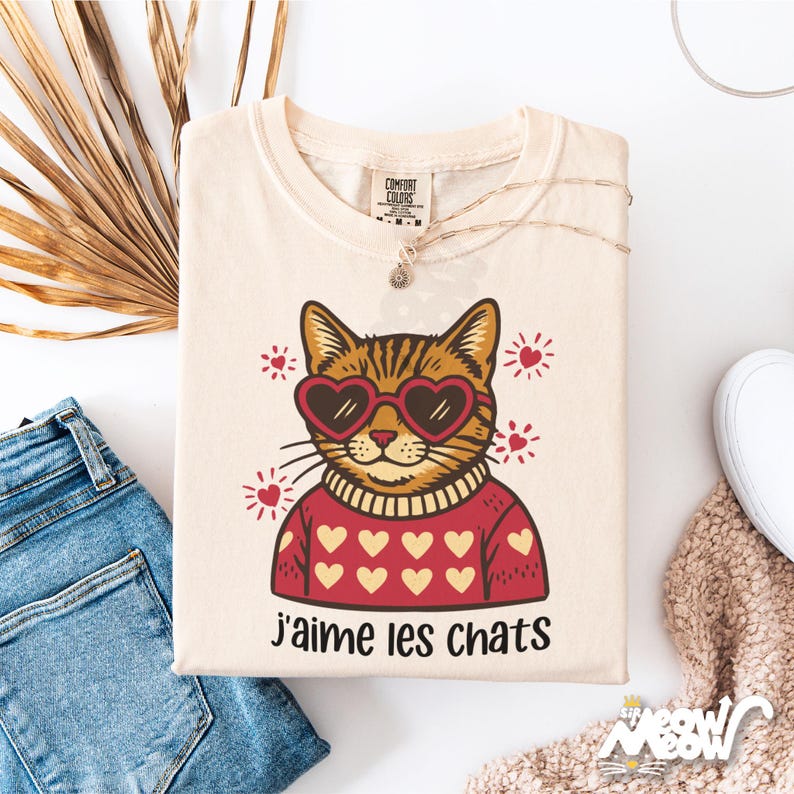 May include: Cream-colored t-shirt featuring a cartoon cat wearing heart-shaped sunglasses and a red sweater with white hearts. The French text "J'aime les chats" is printed below the cat. The shirt is part of a cat-themed apparel collection.