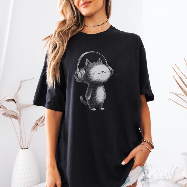 May include: Black t-shirt featuring a grayscale illustration of a cat wearing headphones. The cat is standing upright with a smiling expression. The t-shirt is a relaxed fit, with short sleeves.