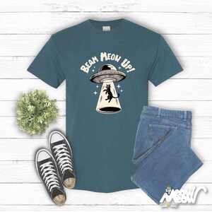 May include: A teal blue t-shirt with a black cat being beamed up by a UFO. The text on the shirt reads "Beam Meow Up!"