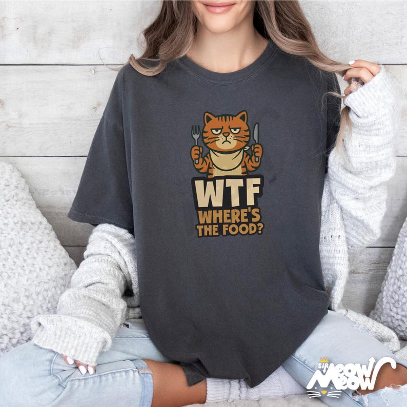 Op de afbeelding: Een donkergrijs T-shirt met een cartoonkat die een vork en mes vasthoudt. De tekst "WTF Where's the Food?" staat onder de kat.