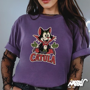 May include: A purple t-shirt featuring a cartoon cat dressed as a vampire with the text "CATULA" in a red and white font. The shirt has rolled-up sleeves and a relaxed fit. The Meow logo is in the bottom right corner.