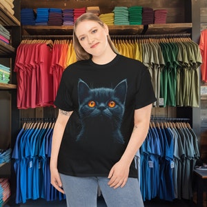 May include: Black t-shirt featuring a detailed illustration of a black cat with bright orange eyes. The cat's fur is depicted with shading, giving it a realistic appearance. The t-shirt is displayed on a person in a clothing store.