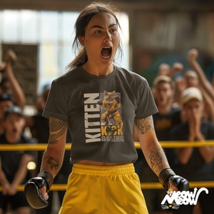 Op de afbeelding: Een vrouw draagt een grijs T-shirt met een afbeelding van een kitten dat boksen. Het shirt zegt "Kitten Kick Boxing". Ze draagt gele shorts en zwarte bokshandschoenen. Ze staat in een boksring met een menigte mensen die kijken.