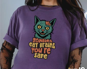 Może przedstawiać: Fioletowy T-shirt z grafiką kreskówkowego zombie kota i tekstem "Zombies eat brains you're safe".