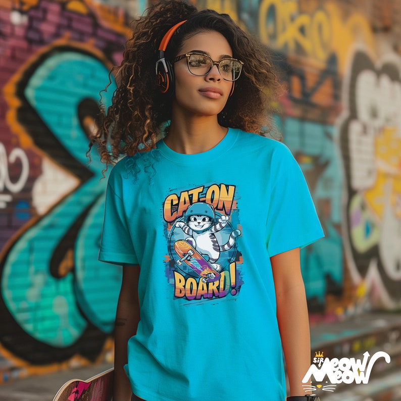 May include: A turquoise blue t-shirt with a graphic of a cat wearing a helmet and riding a skateboard. The graphic has the text "Cat on Board!" in a colorful, pixelated font.