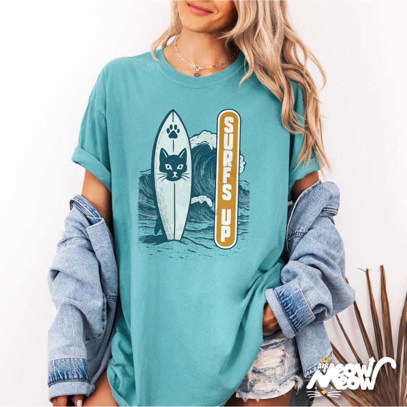 Puede incluir: Camiseta de color turquesa con un dise&ntilde;o de gato surfeando. El gr&aacute;fico muestra un gato en una tabla de surf frente a una ola, con la frase "Surfs Up" impresa verticalmente. La tabla de surf tiene una huella de pata y una cara de gato. La camiseta tiene un corte holgado.