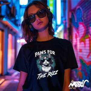 K&ouml;nnte beinhalten: Ein schwarzes T-Shirt mit einem Aufdruck eines Katzen mit Sonnenbrille und Kopfh&ouml;rern. Der Text "PAWS FOR THE RIZZ" ist auf dem Shirt gedruckt.