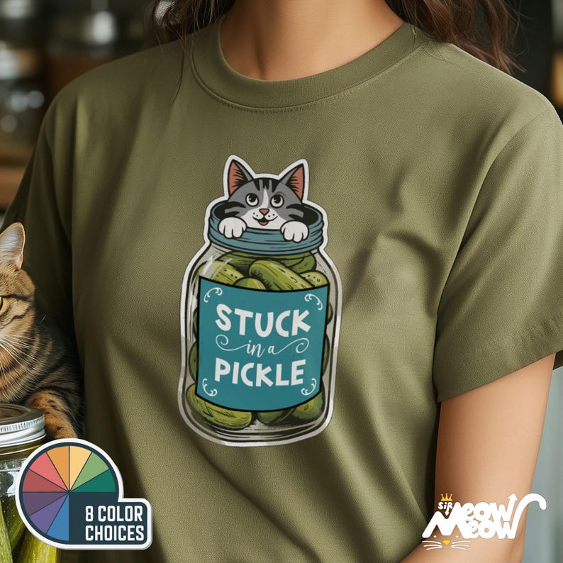 Pu&ograve; includere: Una maglietta verde con una grafica di un gatto bloccato in un barattolo di sottaceti. Il barattolo &egrave; pieno di sottaceti verdi e il gatto sporge dalla parte superiore. Il testo "Stuck in a Pickle" &egrave; stampato sul barattolo.