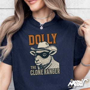 Puede incluir: Camiseta azul marino con la inscripci&oacute;n "DOLLY" en naranja sobre una oveja con sombrero de vaquero y m&aacute;scara. Debajo de la oveja, el texto "THE CLONE RANGER" en color beige. La camiseta tiene cuello redondo.