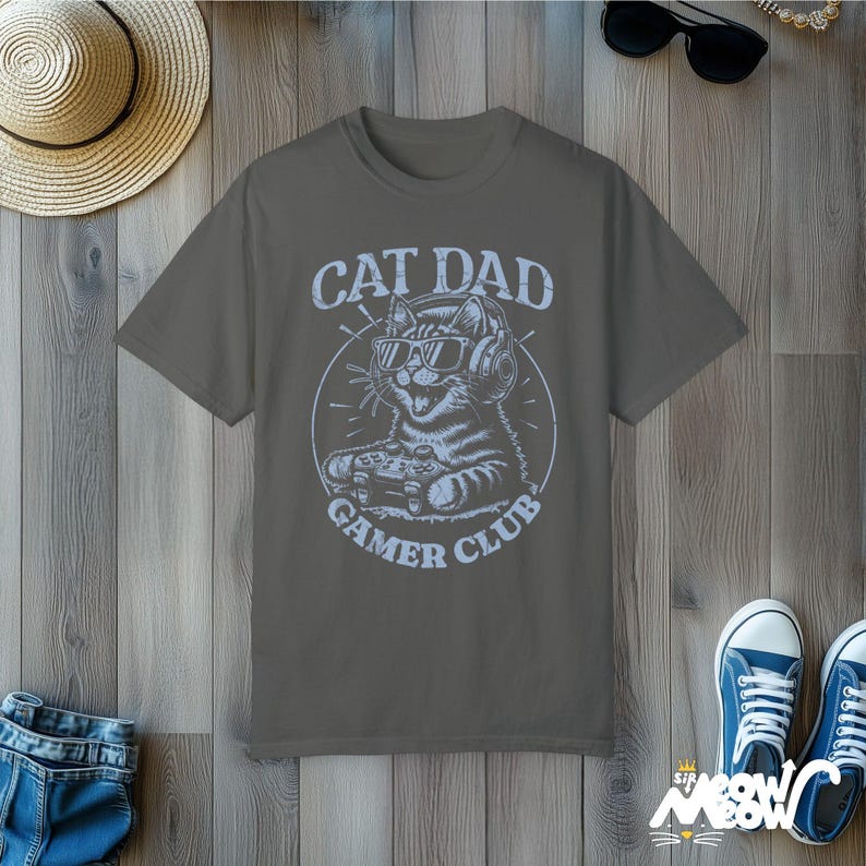 Cat Parent Gamer Club T-Shirts. The tee shows a cat holding a gaming controller. Above the image is the text "Cat Dad" and below the image is the text "Gamer Club".
The t-shirt shown is in the color "Pepper" and is also available in a "Cat Mom" version.