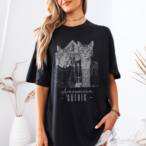 May include: Black t-shirt with a graphic of two cats styled after the American Gothic painting. The cats are dressed in period clothing and hold a pitchfork. The text "Ameowican GOTHIC" is printed below the image.