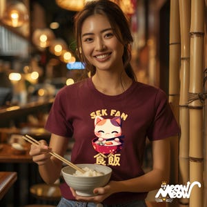 May include: A maroon t-shirt with a cartoon cat eating from a bowl of food. The text "SEK FAN" is above the cat and the text "食飯" is below the cat. The cat has a white and orange face and a pink nose.
