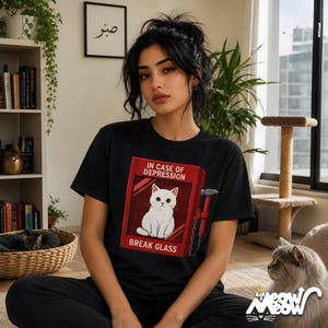 May include: Black t-shirt featuring a red graphic of a fire alarm box with a white cat inside and the text "IN CASE OF DEPRESSION BREAK GLASS." The shirt has a crew neck and short sleeves. The image also includes a cat.