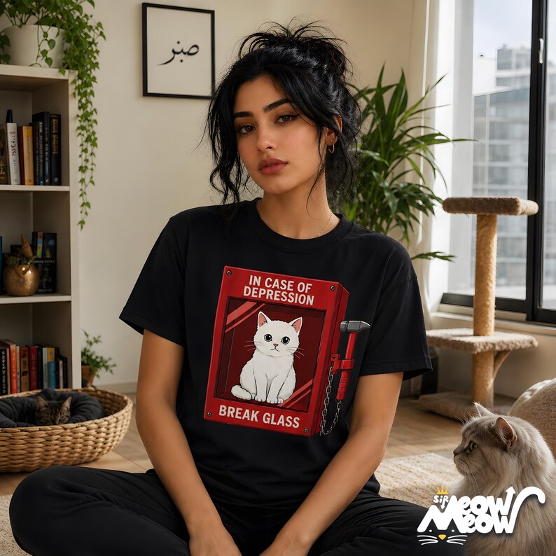May include: Black t-shirt featuring a red graphic of a fire alarm box with a white cat inside and the text "IN CASE OF DEPRESSION BREAK GLASS." The shirt has a crew neck and short sleeves. The image also includes a cat.