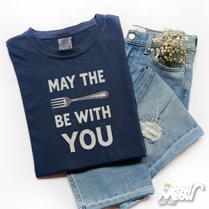 Peut inclure: Un t-shirt bleu marine avec le texte "MAY THE FORK BE WITH YOU" et un dessin de fourchette, associ&eacute; &agrave; un short et un jean en denim bleu clair. Un petit bouquet de fleurs blanches est dans la poche du jean.