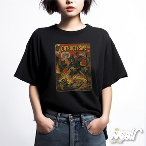 Pu&ograve; includere: T-shirt nera con grafica a fumetti con il titolo "CAT-ACLYSM!" e un gatto supereroe in azione. Il design include i testi "SKRRRAK!" e "CAT-TANA SAVES THE FANG FURY!"