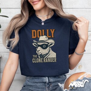 Puede incluir: Camiseta azul marino con un dise&ntilde;o de una oveja con sombrero de vaquero y gafas de sol, con el texto "DOLLY THE CLONE RANGER" en marr&oacute;n y blanco. La camiseta tiene cuello redondo y mangas cortas.