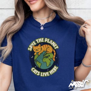 以下が含まれることがあります： 地球で眠る猫のグラフィックがプリントされたネイビーブルーのTシャツ。Tシャツには「Save the Planet Cats Live Here」の文字がプリントされています。