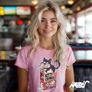 Może przedstawiać: R&oacute;żowy T-shirt z grafiką kresk&oacute;wkowego kota na pudełku płatk&oacute;w śniadaniowych Kitty Crunch. Pudełko ma napis "Kitty Crunch" i "Meow!" na nim.