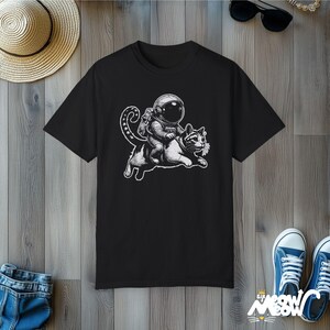 Black Comfort Colors 1717 unisex T-shirt with a humorous white graphic of an astronaut riding atop a playful cat, combining outer space and cat lover themes.