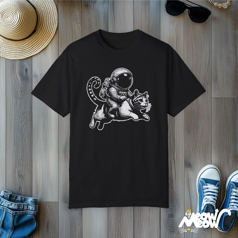 Black Comfort Colors 1717 unisex T-shirt with a humorous white graphic of an astronaut riding atop a playful cat, combining outer space and cat lover themes.