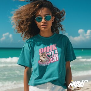 K&ouml;nnte beinhalten: T&uuml;rkisfarbenes T-Shirt mit einem Aufdruck einer Katze auf einem rosa Flamingo-Schwimmring, dem Text "PINK FLA MEOW GO". Das Shirt hat eine lockere Passform und kurze &Auml;rmel. Das Bild ist vor einem Strand und blauem Himmel aufgenommen.