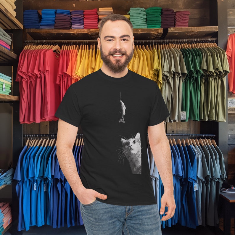 May include: A black t-shirt with a white cat looking up at a fish on a fishing line. The cat is in the lower right corner of the shirt. The fish is in the upper center of the shirt.