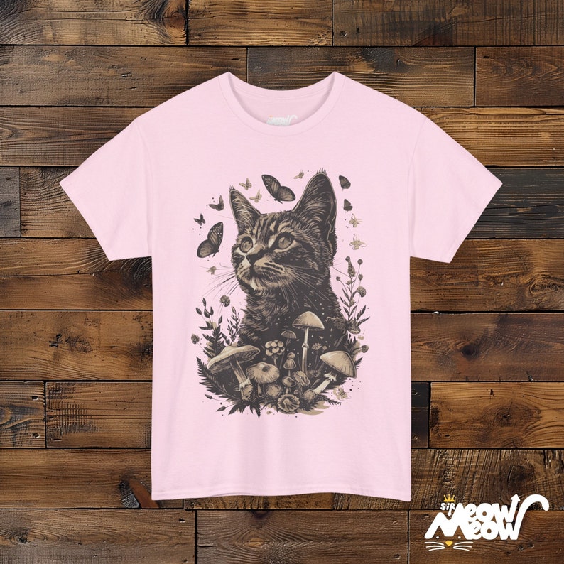 Vintage style t-shirt print of a cat in the woodlands surrounded by wildflowers, mushrooms and butterflies. Light pink tee.