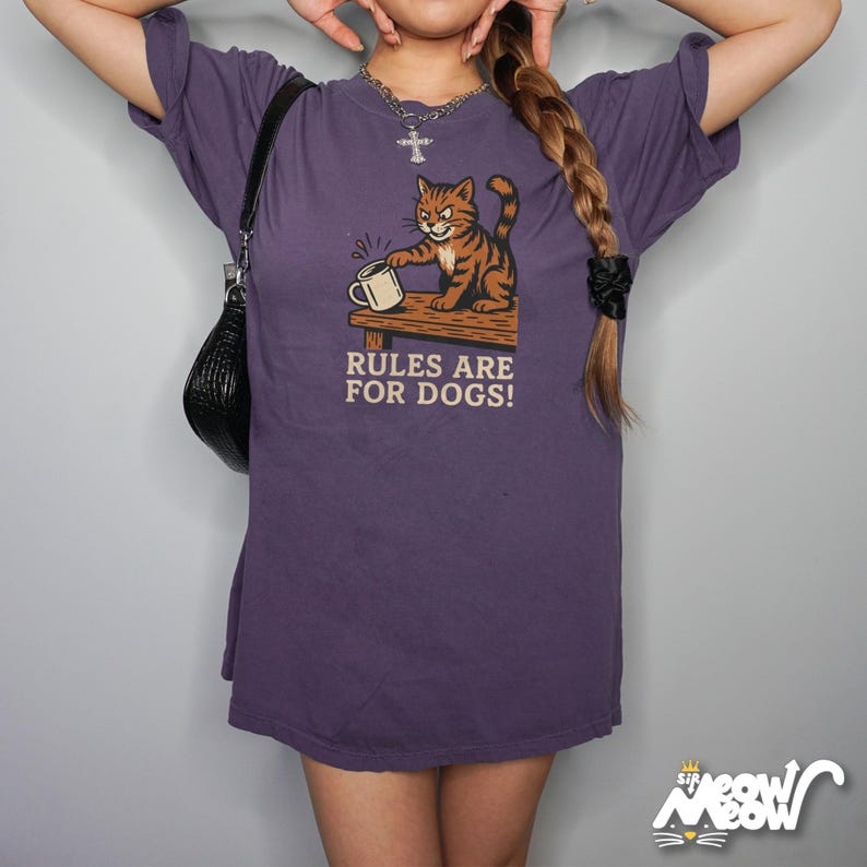 May include: A purple t-shirt with a graphic of a cartoon cat holding a mug, with the text "RULES ARE FOR DOGS!". The cat is orange and brown. The shirt is a loose fit.