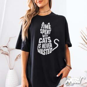 May include: Black t-shirt with a white cat silhouette design. The cat-shaped design contains the text "TIME SPENT WITH CATS IS NEVER WASTED". The shirt has short sleeves and a crew neck.