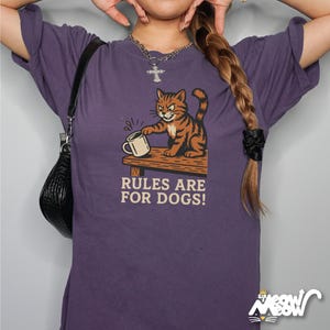 May include: Purple t-shirt featuring a cartoon cat with orange and brown stripes, holding a mug on a wooden table. The text "RULES ARE FOR DOGS!" is below the cat illustration. The shirt has short sleeves.