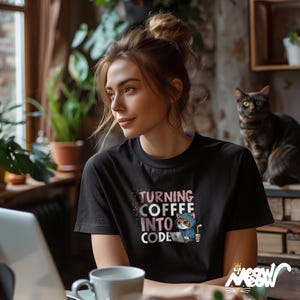 May include: A black t-shirt featuring a pink and white graphic of a cat wearing a blue hoodie and holding a coffee cup. The text on the shirt reads "Turning Coffee Into Code".