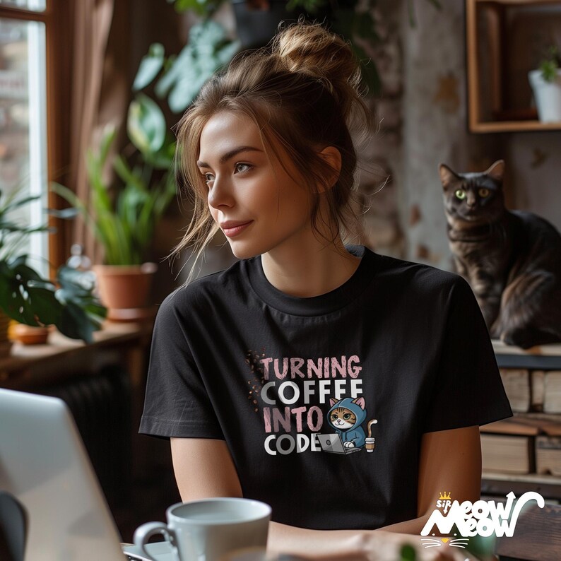 May include: A black t-shirt featuring a pink and white graphic of a cat wearing a blue hoodie and holding a coffee cup. The text on the shirt reads "Turning Coffee Into Code".