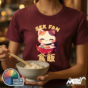 May include: A maroon t-shirt with a cartoon cat eating ramen. The cat has white fur with orange and black markings. The text on the shirt reads "Sek Fan" and "食飯".