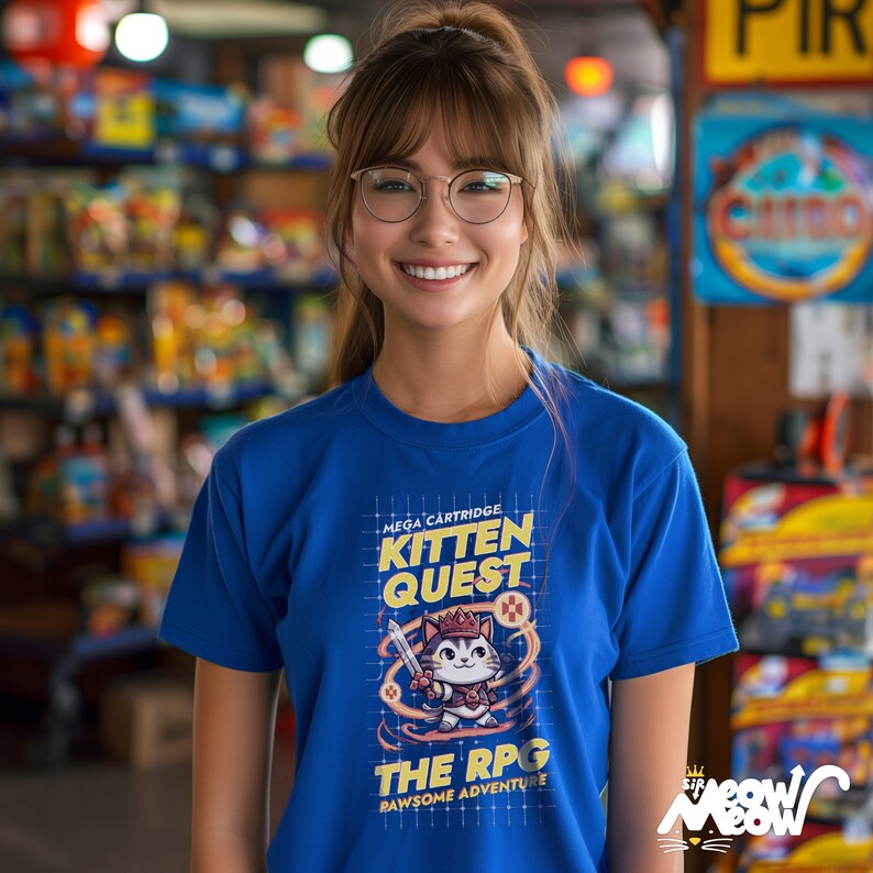 May include: A woman wearing a blue t-shirt with a graphic of a cat in a crown holding a sword. The graphic says "Mega Cartridge Kitten Quest The RPG Pawsome Adventure".