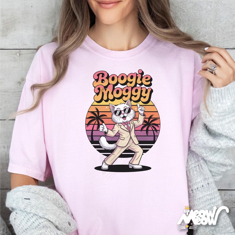 May include: Light pink t-shirt featuring a cartoon cat in a beige suit and sunglasses, dancing in front of a sunset with palm trees. The text "Boogie Moggy" is above the cat in a retro font.