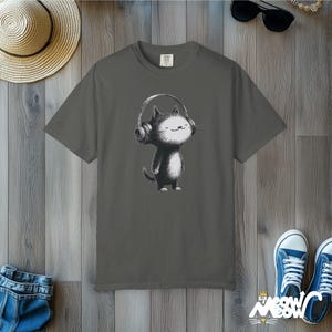 May include: Gray t-shirt featuring a black and white cartoon cat wearing headphones. The cat has a happy expression. The shirt is displayed on a wooden surface with a straw hat, sunglasses, and blue sneakers.