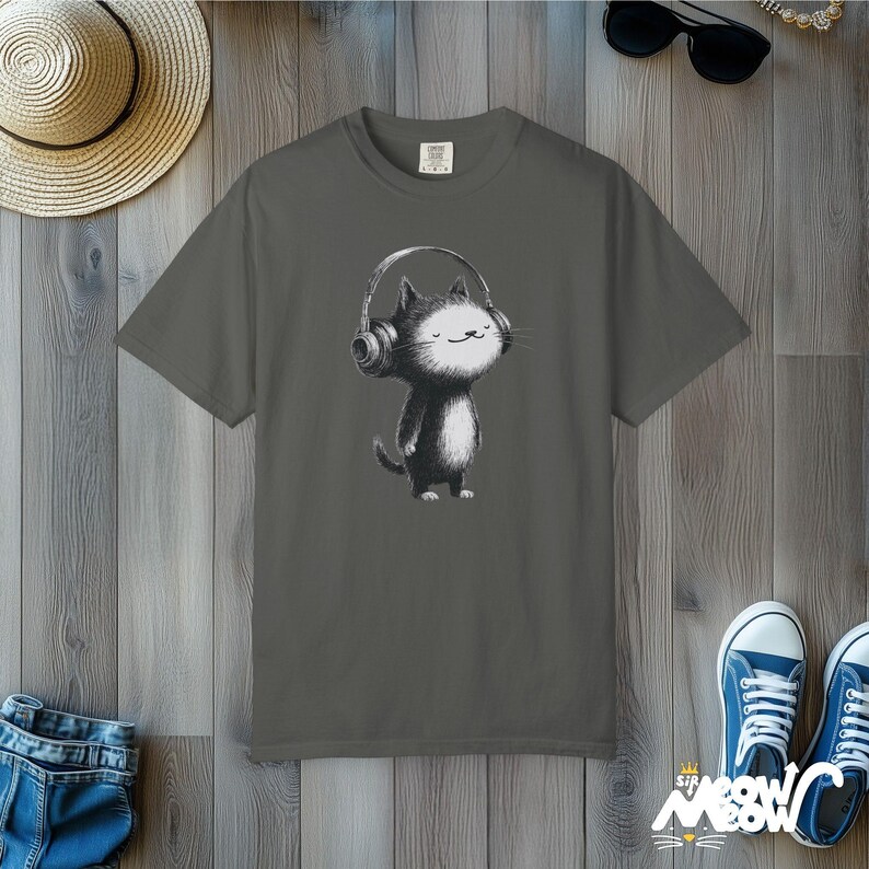 May include: Gray t-shirt featuring a black and white cartoon cat wearing headphones. The cat has a happy expression. The shirt is displayed on a wooden surface with a straw hat, sunglasses, and blue sneakers.