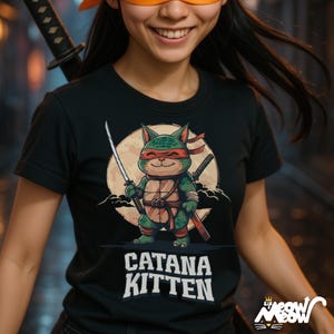May include: Black t-shirt featuring a cartoon cat ninja with swords, a red headband, and a turtle shell design. The shirt has the text "Catana Kitten" in white block letters. The design is set against a full moon background.