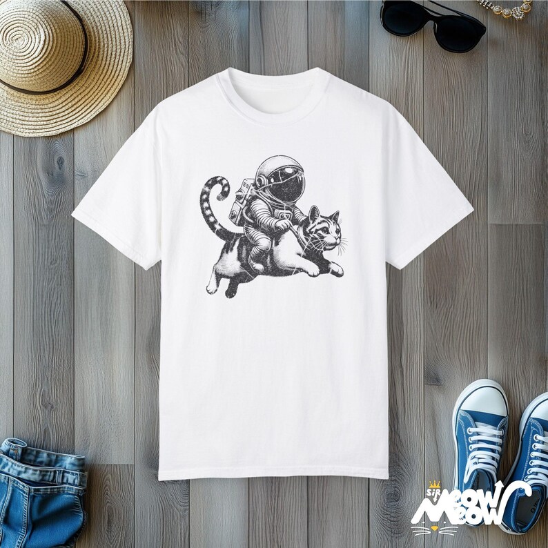White Comfort Colors 1717 unisex T-shirt with a humorous white graphic of an astronaut riding atop a playful cat, combining outer space and cat lover themes.