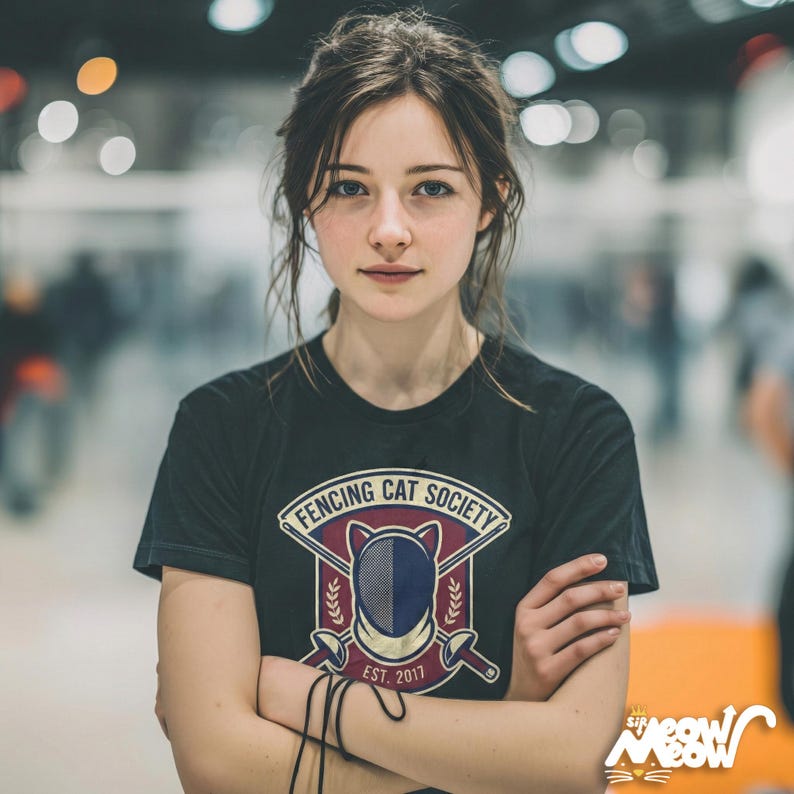 May include: A black t-shirt with the words "FENCING CAT SOCIETY" in a circular design, featuring a cat silhouette and fencing equipment. The shirt also has the text "EST. 2011". The person is wearing the shirt.