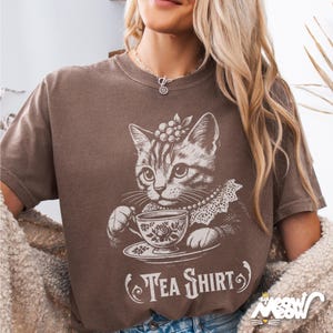 May include: A brown t-shirt with a graphic of a cat drinking tea from a teacup. The text "Tea Shirt" is printed below the cat.