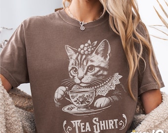 May include: A brown t-shirt with a graphic of a cat drinking tea from a teacup. The text "Tea Shirt" is printed below the cat.