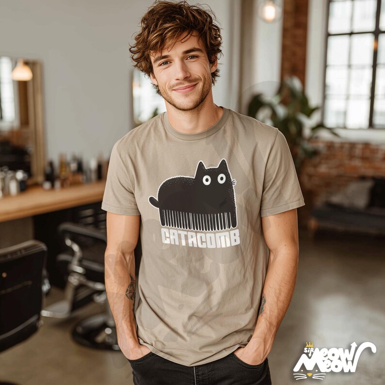 May include: A tan t-shirt featuring a black cat design with a comb for a body and the text "CATACOMB" in white. The shirt has short sleeves and a crew neck. The design is a playful take on a cat and comb.
