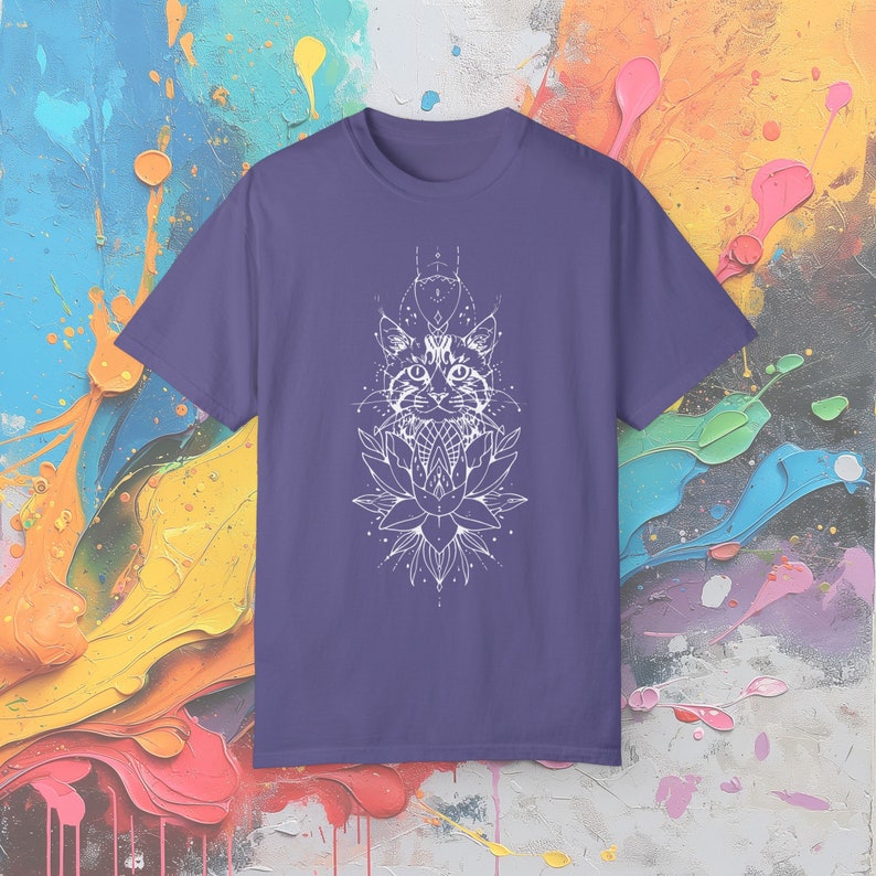 May include: A purple t-shirt featuring a white graphic of a cat's face, a lotus flower, and geometric designs. The cat's face is centered above the lotus flower. The design is detailed and intricate, with fine lines and shading.