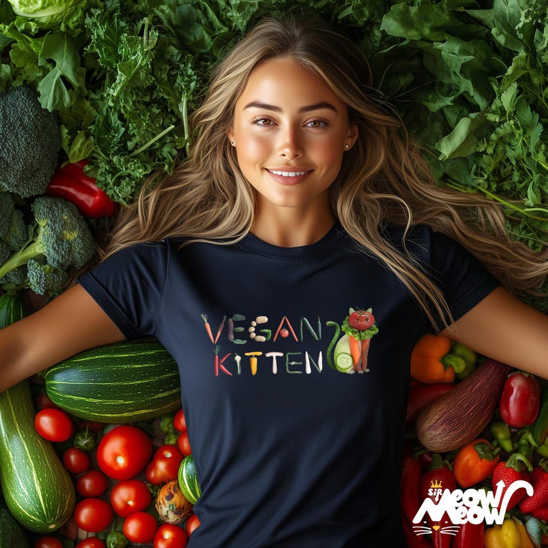 Puede incluir: Una mujer lleva una camiseta azul oscuro con el texto "VEGAN? KITTEN" en letras de colores hechas de verduras. El texto est&aacute; rodeado de una variedad de verduras, incluyendo tomates, calabacines y br&oacute;coli.