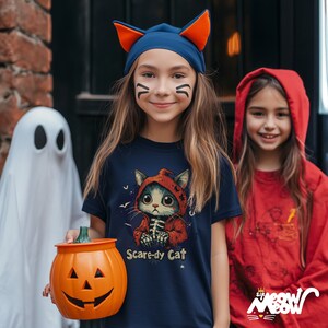 Puede incluir: Dos ni&ntilde;as vestidas con disfraces de Halloween. Una lleva una camiseta azul marino con un gr&aacute;fico de "Scare-dy Cat" y una diadema con orejas de gato. La otra lleva una sudadera con capucha roja. Se sostiene un cubo de calabaza naranja tallada en primer plano.