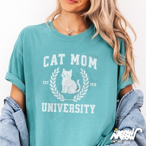 May include: A teal blue t-shirt with the text "Cat Mom University" in white. The text is in a vintage collegiate style font. A white cat illustration is in the center of the text.