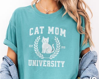 May include: A teal blue t-shirt with the text "Cat Mom University" in white. The text is in a vintage collegiate style font. A white cat illustration is in the center of the text.
